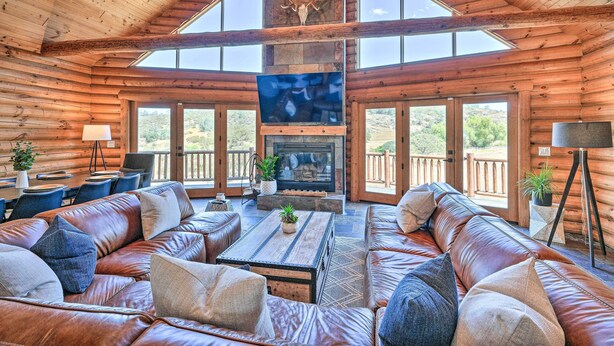 A log cabin interior with a large wooden ceiling, a stone fireplace, leather sofas, and large windows.