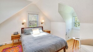 A bedroom with a sloped ceiling, a bed with striped bedding, two bedside lamps, a nightstand, a small table, and a window with a view. - Quaint Woodstock Escape - 2 Mi to Tinker St! (West Hurley)