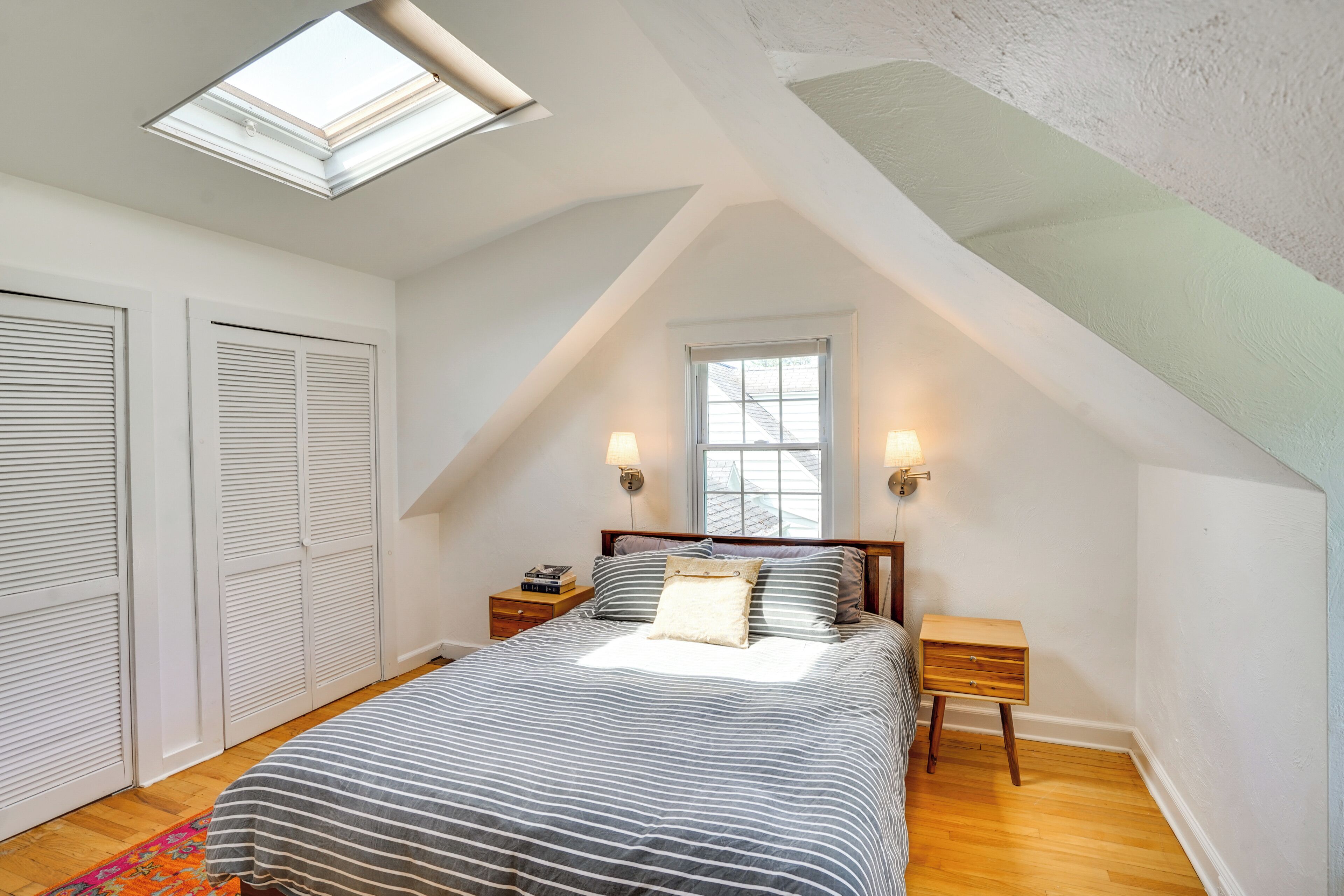 A bedroom with a sloped ceiling, a bed with striped bedding, two bedside tables with lamps, and a window with blinds.