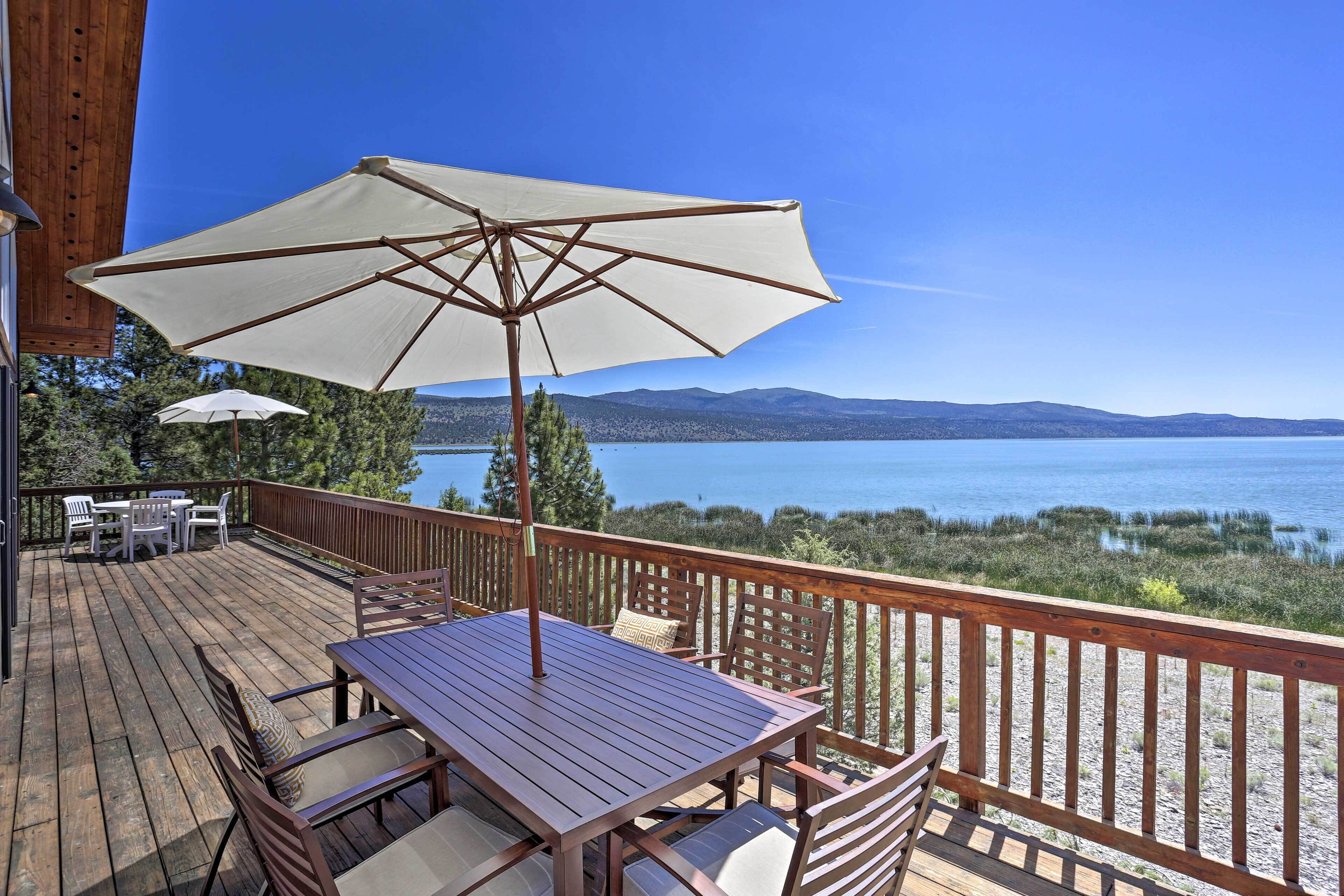 Une terrasse en bois avec une table et des chaises, un parasol, et une vue sur un lac et des montagnes.