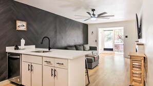 Una cocina moderna con una isla blanca, grifo negro y armarios de madera. Hay un ventilador de techo, una pequeña mesa de madera y vistas al exterior a través de una puerta corrediza.
