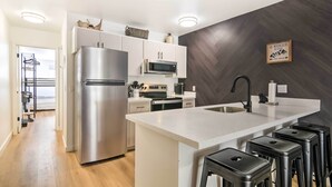 Una cocina moderna con un refrigerador de acero inoxidable, gabinetes blancos, un microondas y un lavamanos. Hay taburetes negros en la isla y un cartel decorativo en la pared.