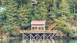 A wooden cabin on stilts over a body of water, surrounded by dense forest. - Serene Seldovia Cabin w/ Deck, Grill & Views! (Seldovia)