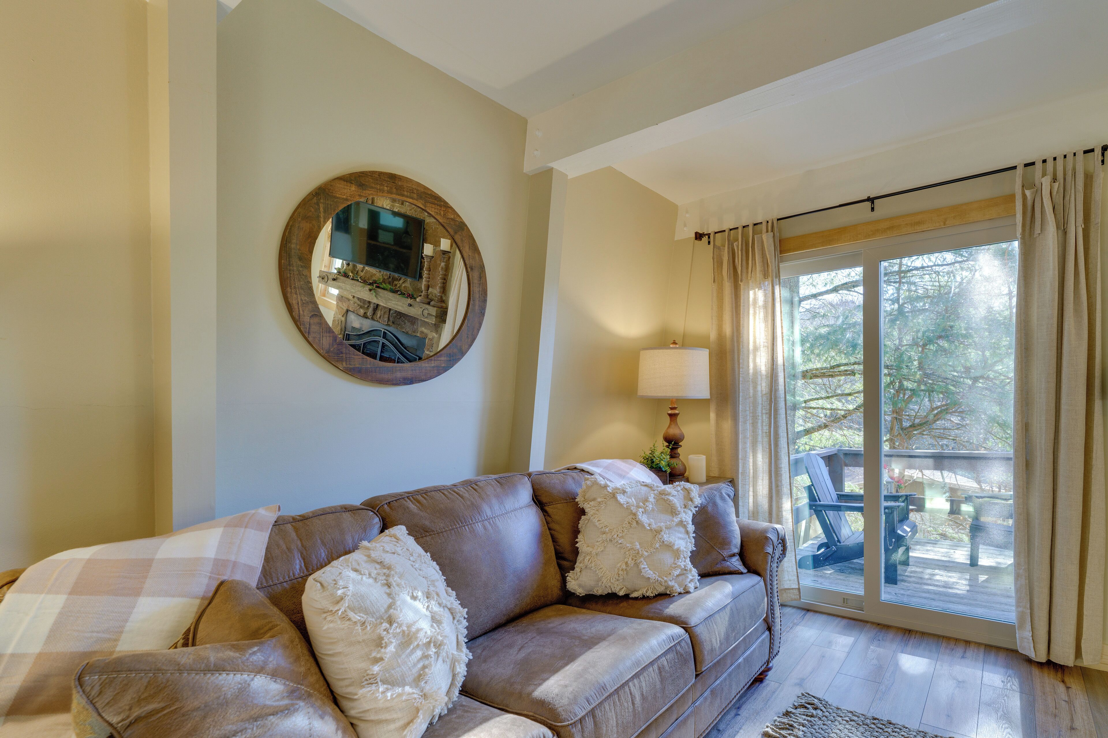 A living room with a brown leather sofa, a round mirror on the wall, a lamp, and a view through a glass door to an outdoor deck.