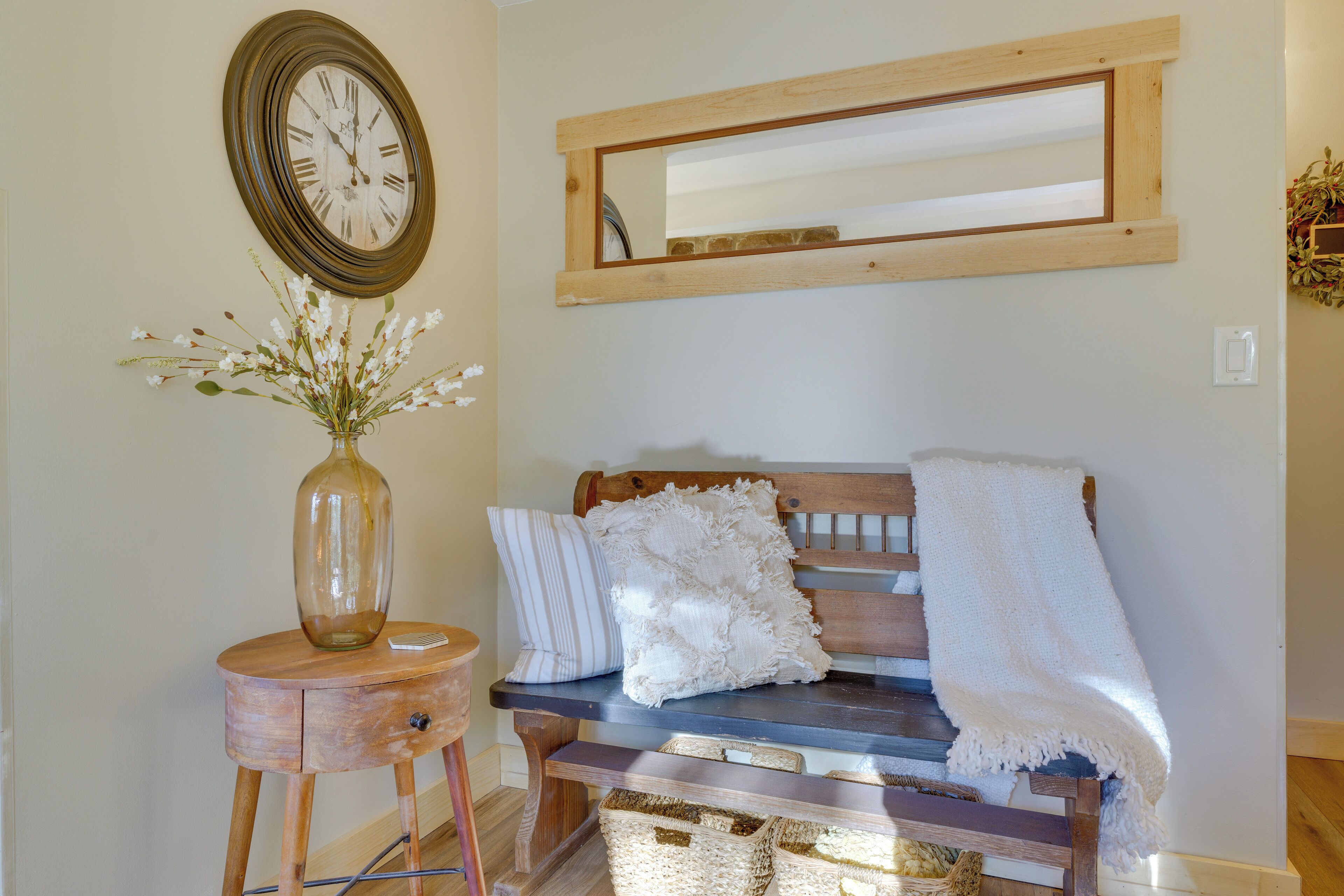A wooden bench with a white throw blanket, a mirror, and a vase with flowers on a wooden stool.