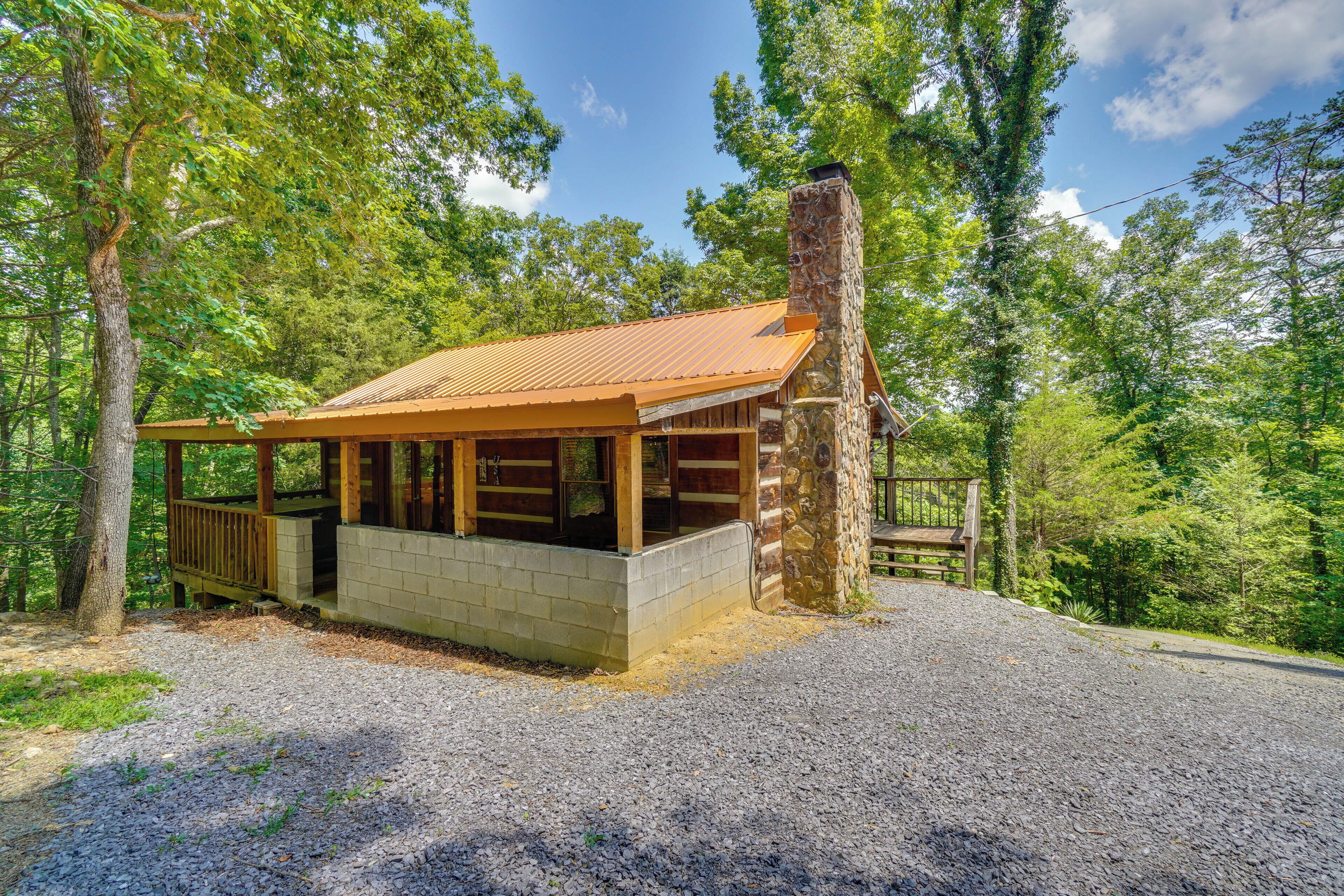 A rustic cabin with a stone chimney, wooden deck, and a gravel driveway surrounded by trees.