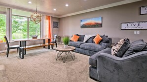 A modern living room with a dining area, grey sofas, a wooden coffee table, and a large window providing a view of greenery.