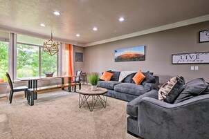 A modern living room with a dining area, grey sofas, a wooden coffee table, and a large window providing a view of greenery.