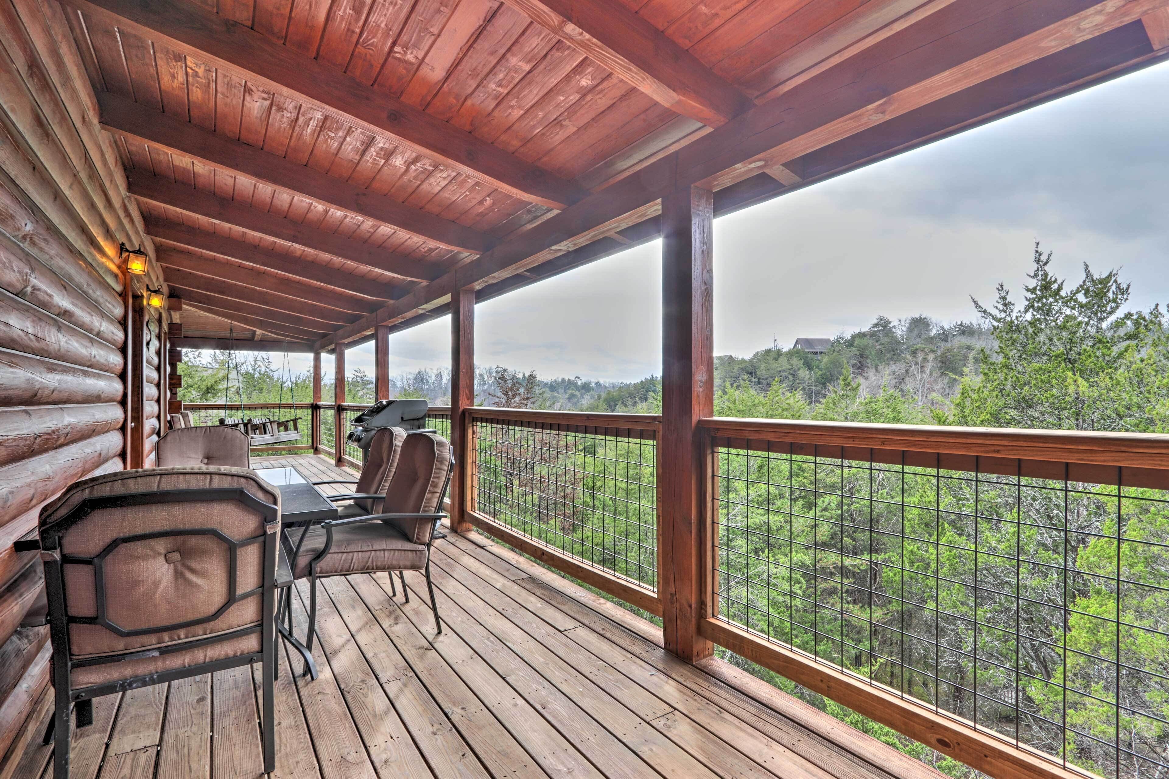 Una terraza techada con piso de madera, una mesa con sillas y vista a un paisaje boscoso.