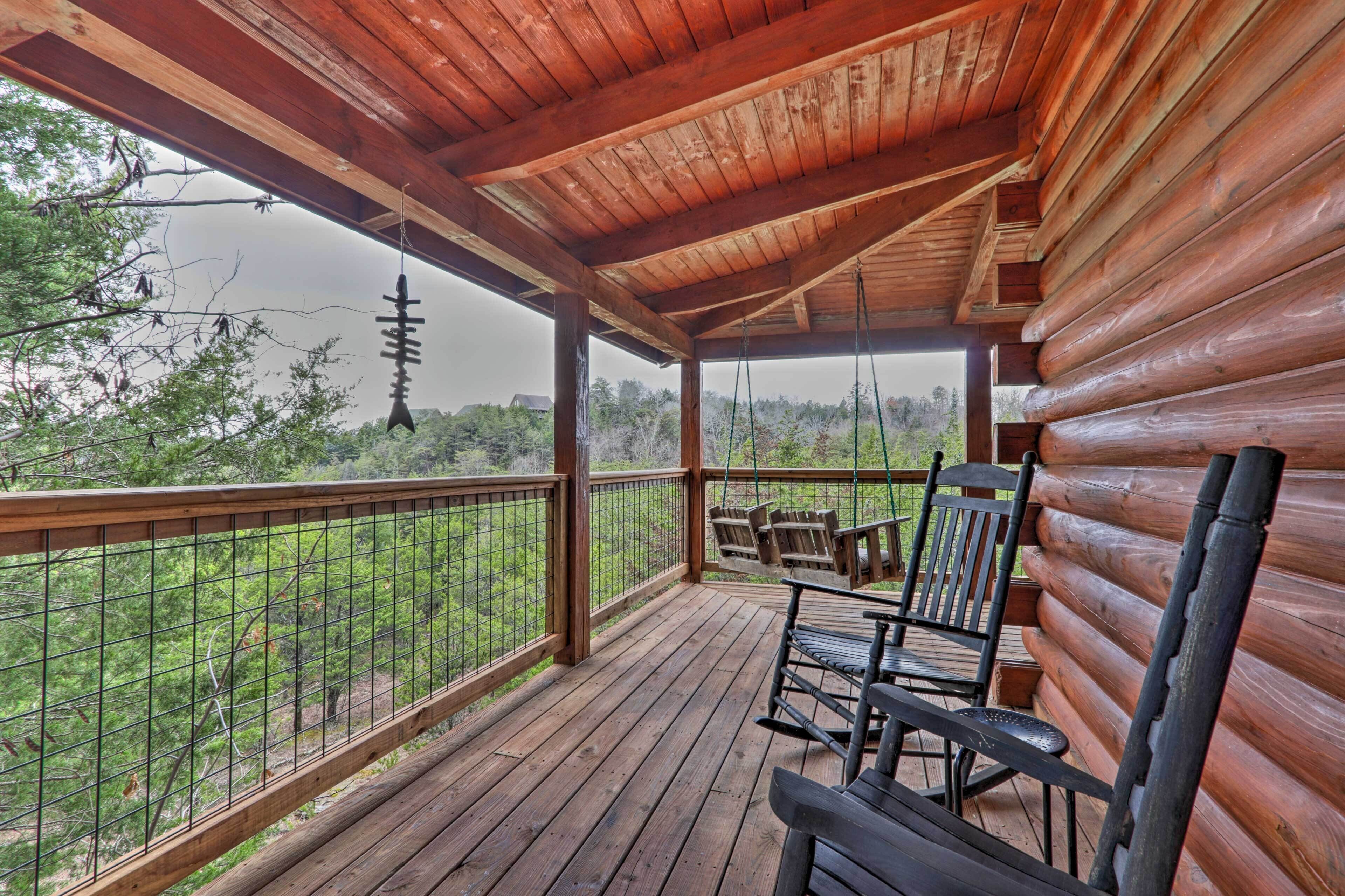 A wooden deck with a covered area, wooden chairs, and a view of the surrounding forest.