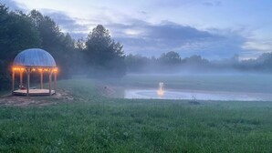 Um gazebo com luzes de festa em um campo gramado com um lago e névoa.