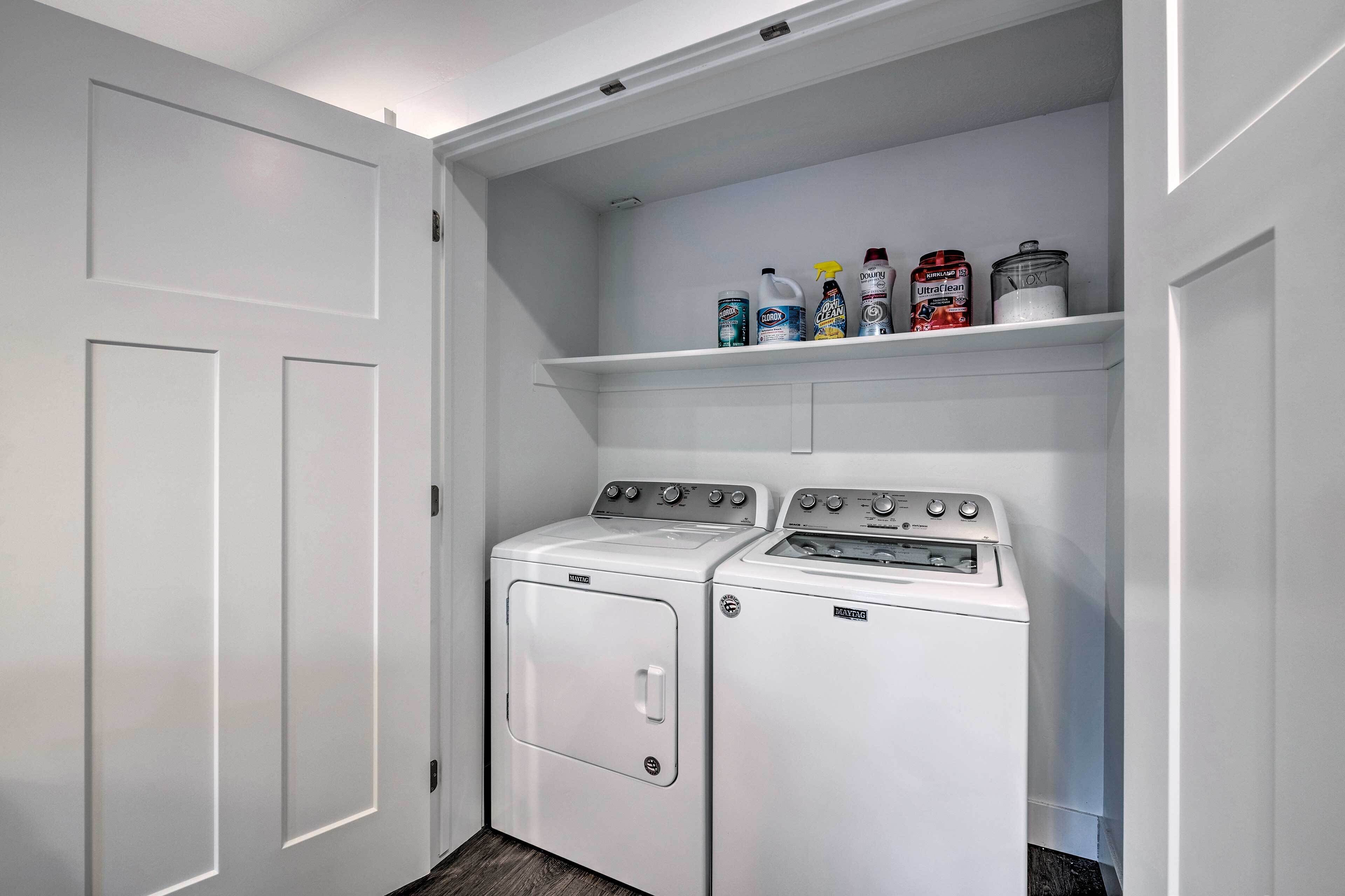 Une buanderie équipée de deux machines à laver et de deux sèche-linge à chargement frontal, tous deux blancs, d’une étagère contenant des produits de nettoyage et d’une porte blanche.