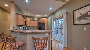 A kitchen with wooden cabinets, a central island with bar stools, a refrigerator, and a window with blinds. - Sevierville Cabin w/ Lake Views & Wraparound Porch (Sevierville)