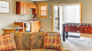 A cozy living room with a sofa, a foosball table, and a kitchen area with wooden cabinets and a window with blinds. - Sevierville Cabin w/ Lake Views & Wraparound Porch (Sevierville)