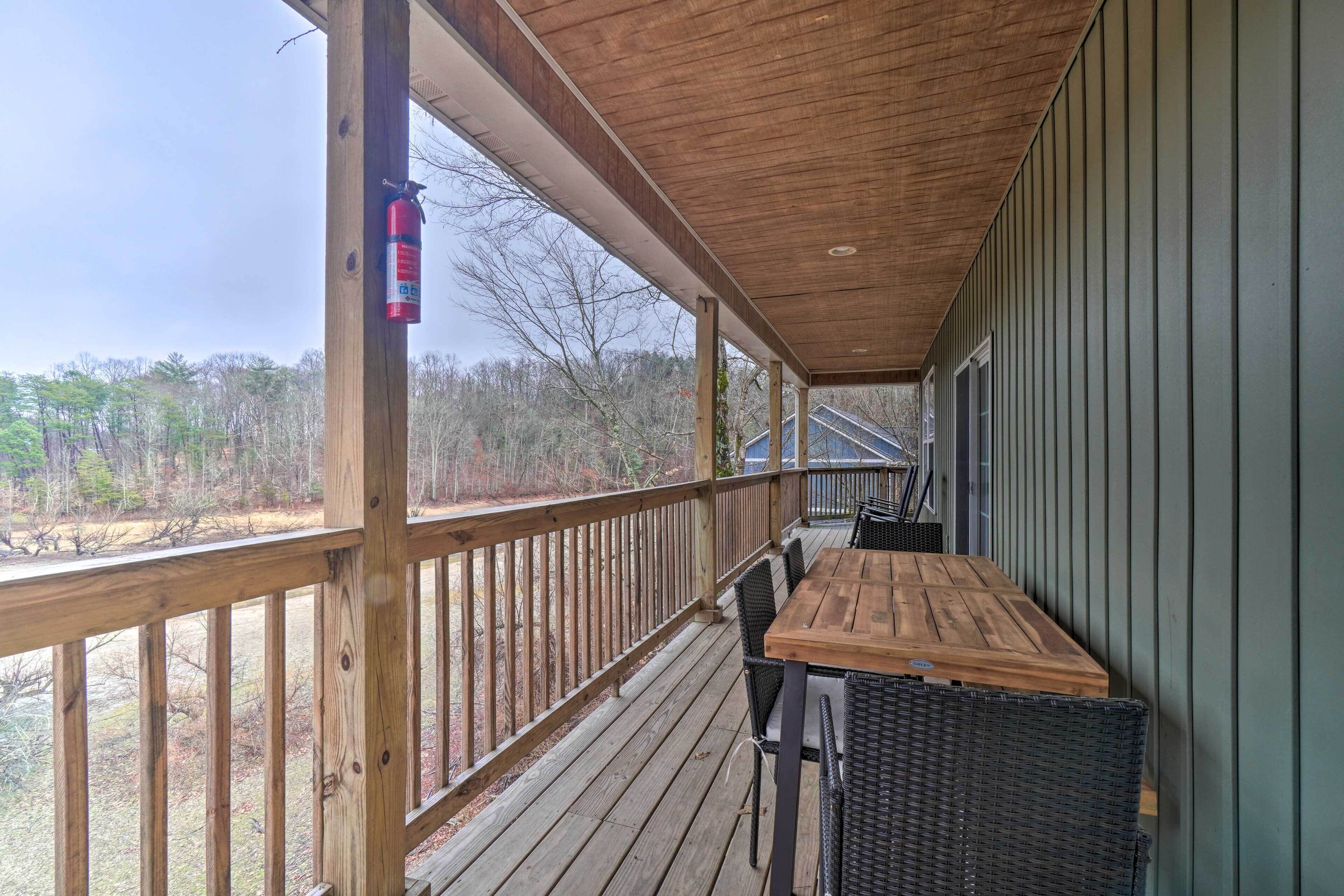 A covered outdoor deck with a wooden table and chairs, a fire extinguisher, and a view of trees and a building in the background.