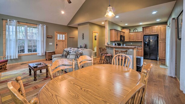 A spacious living area with a wooden dining table, chairs, and a sofa. A kitchen with wooden cabinets and a refrigerator is visible in the background. - Sevierville Cabin w/ Lake Views & Wraparound Porch (Sevierville)