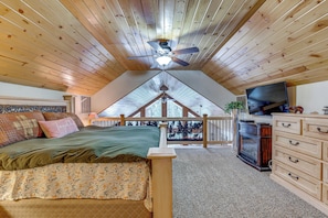 Une chambre avec un plafond en pente, un lit recouvert d’une couvre-lit à motifs floraux, une commode, un téléviseur et un balcon offrant une vue sur les arbres.