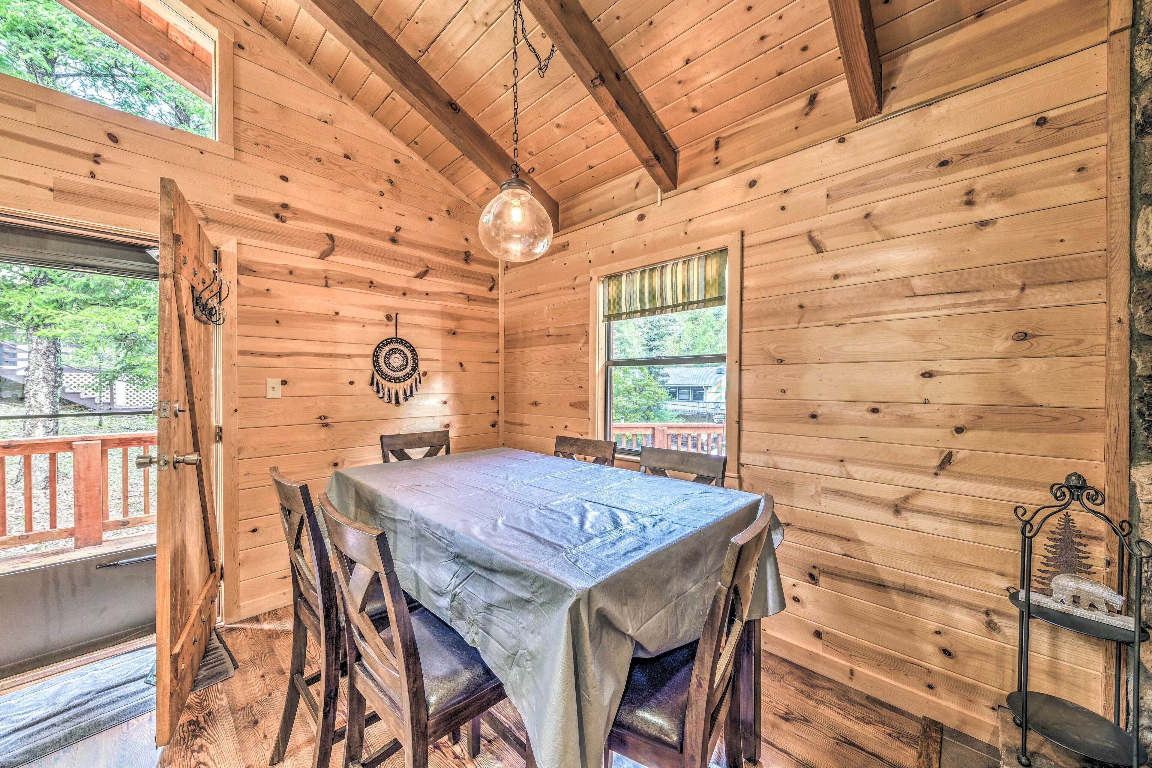 A wooden cabin with a dining table, chairs, a hanging light fixture, and a window with a view of greenery.
