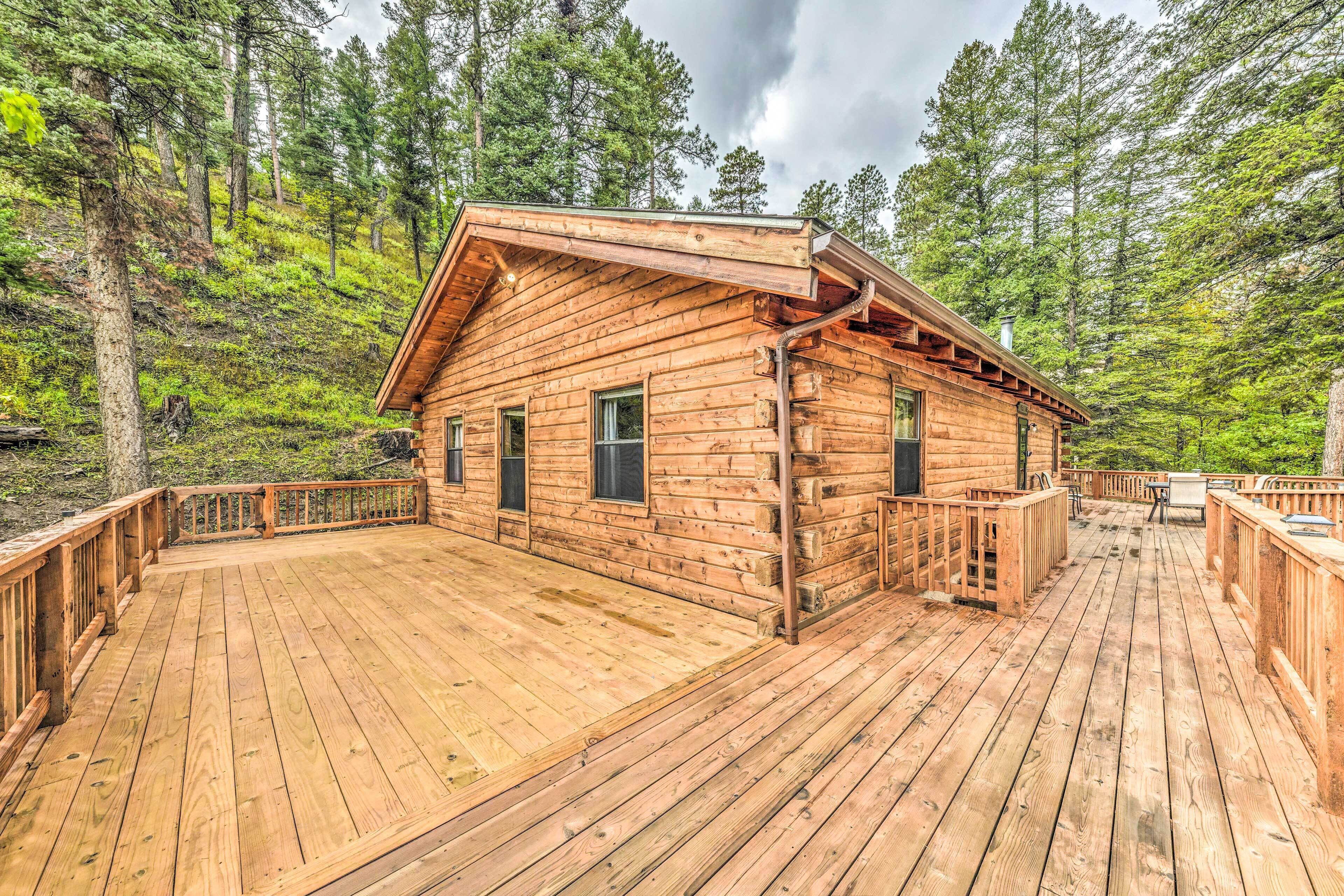 A wooden cabin with a deck, surrounded by forest.
