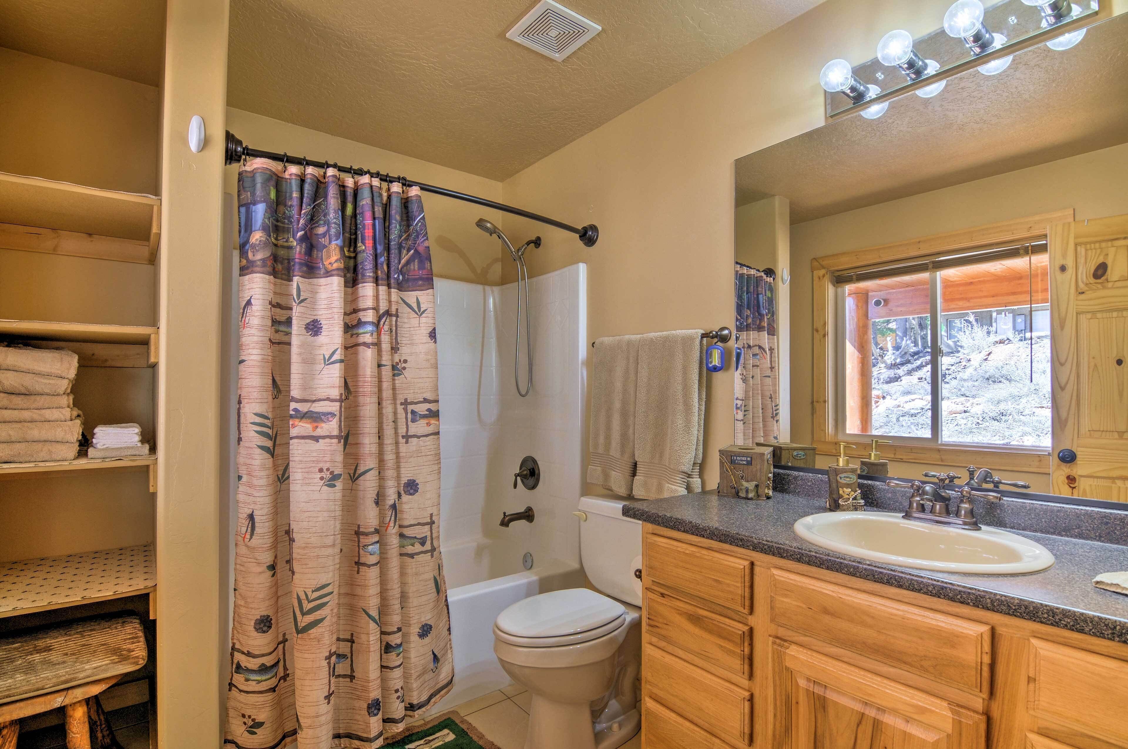 Un baño con bañera, inodoro y lavamanos. Hay una cortina de ducha con estampado floral, un perchero y una ventana con vistas al exterior.