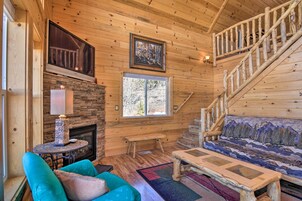 A wooden cabin interior with a fireplace, a sofa, a bench, a staircase, and a large window.