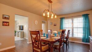 A dining area with a wooden table set for four, surrounded by chairs. There is a chandelier above the table, and a window with curtains to the right. - The Village Home w/ Pool, 12 Mi to Okc! (Oklahoma City)