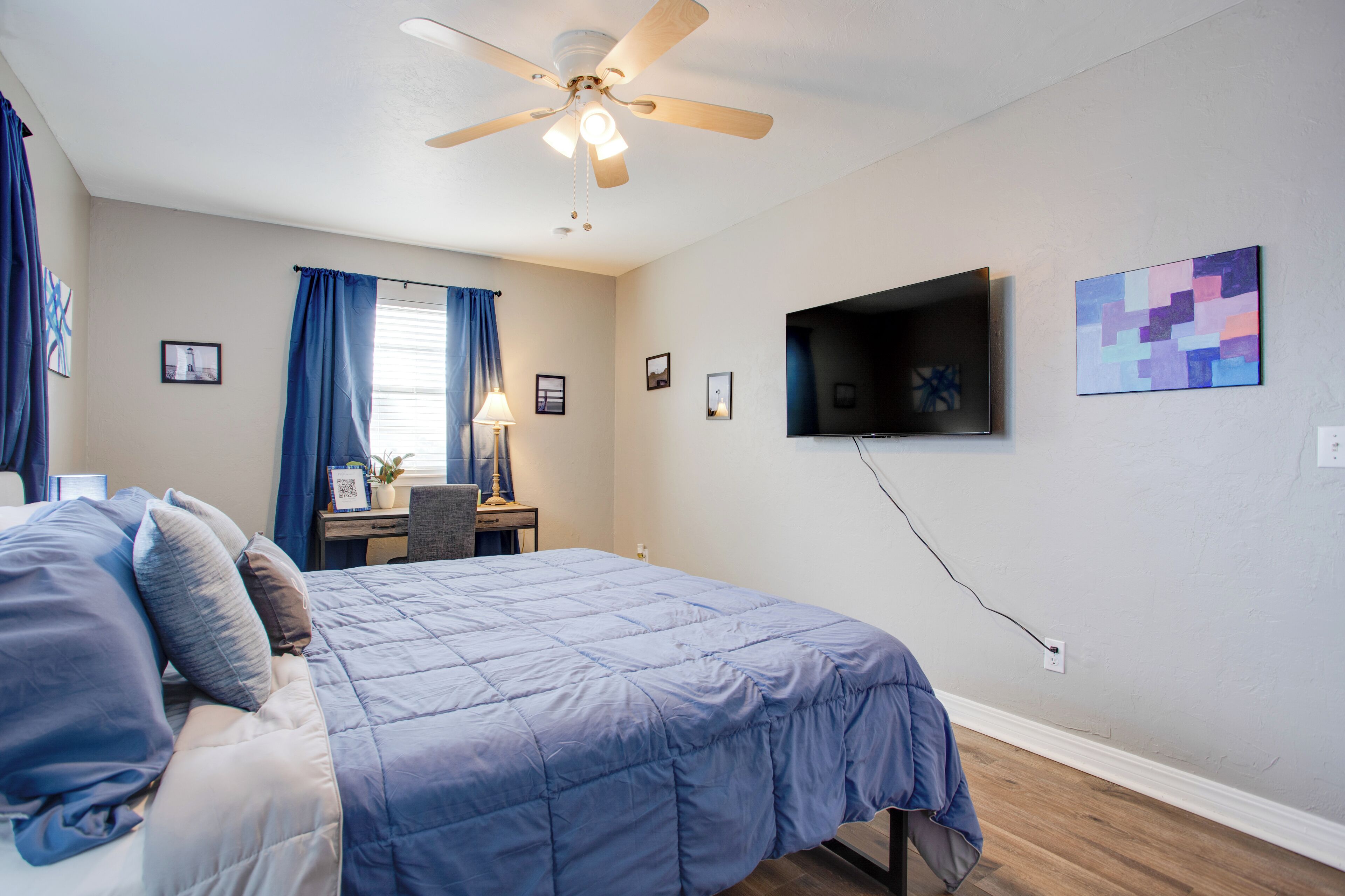 A bedroom with a bed, a desk, a chair, a ceiling fan, a TV mounted on the wall, and a window with curtains.