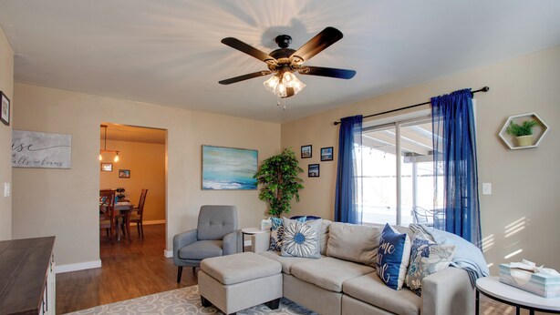 A living room with a ceiling fan, a sofa, an ottoman, a dining area with a table and chairs, and a window with curtains. - The Village Home w/ Pool, 12 Mi to Okc! (Oklahoma City)