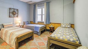 Una habitación con tres camas, una mesita, una lámpara y un ventanal con cortinas.