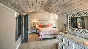 Een slaapkamer met een schuine plafond, een bed met een bedspreide met patroon, een dressoir met lades, een spiegel en een nachtkastje met een lamp.