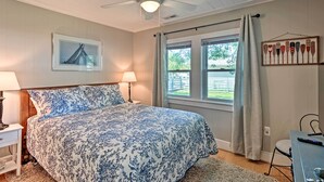 Un dormitorio con cama, ventilador de techo, ventana con cortinas, un cuadro en la pared y un escritorio con silla.