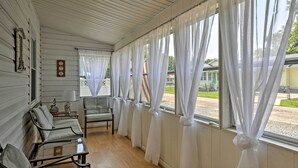 Una habitación con una fila de sillas, una mesa y ventanas grandes con cortinas blancas.
