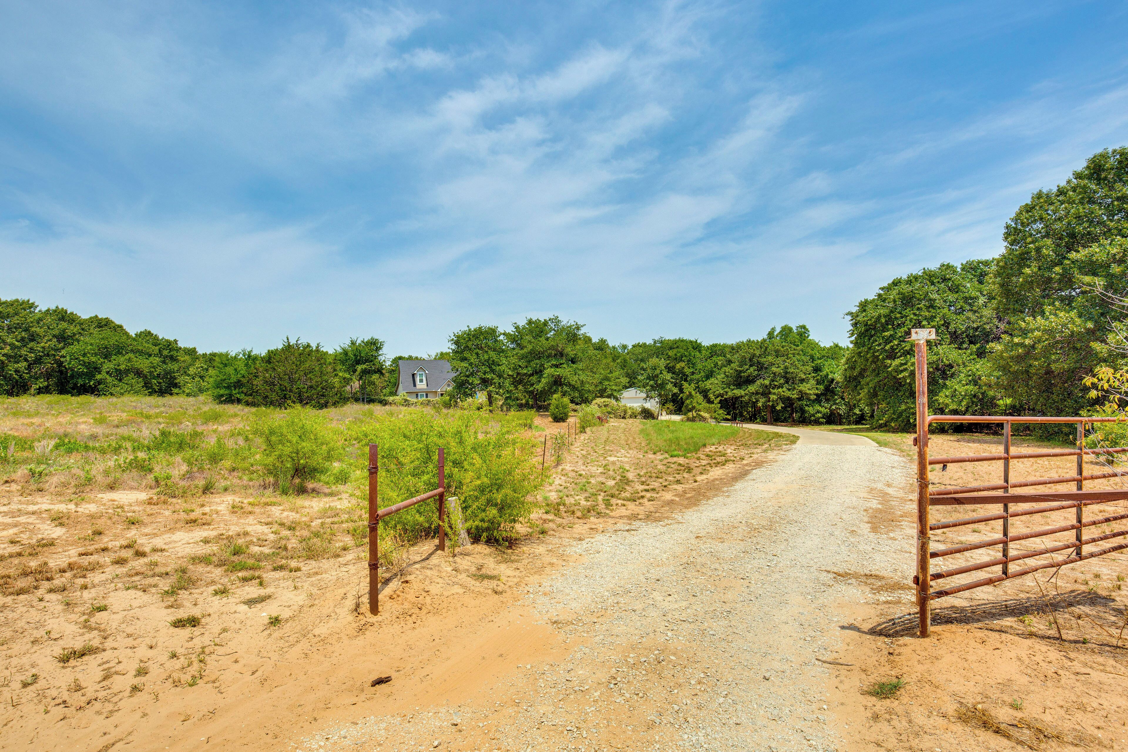 A rural dirt road leading to a gated area with a house in the background.