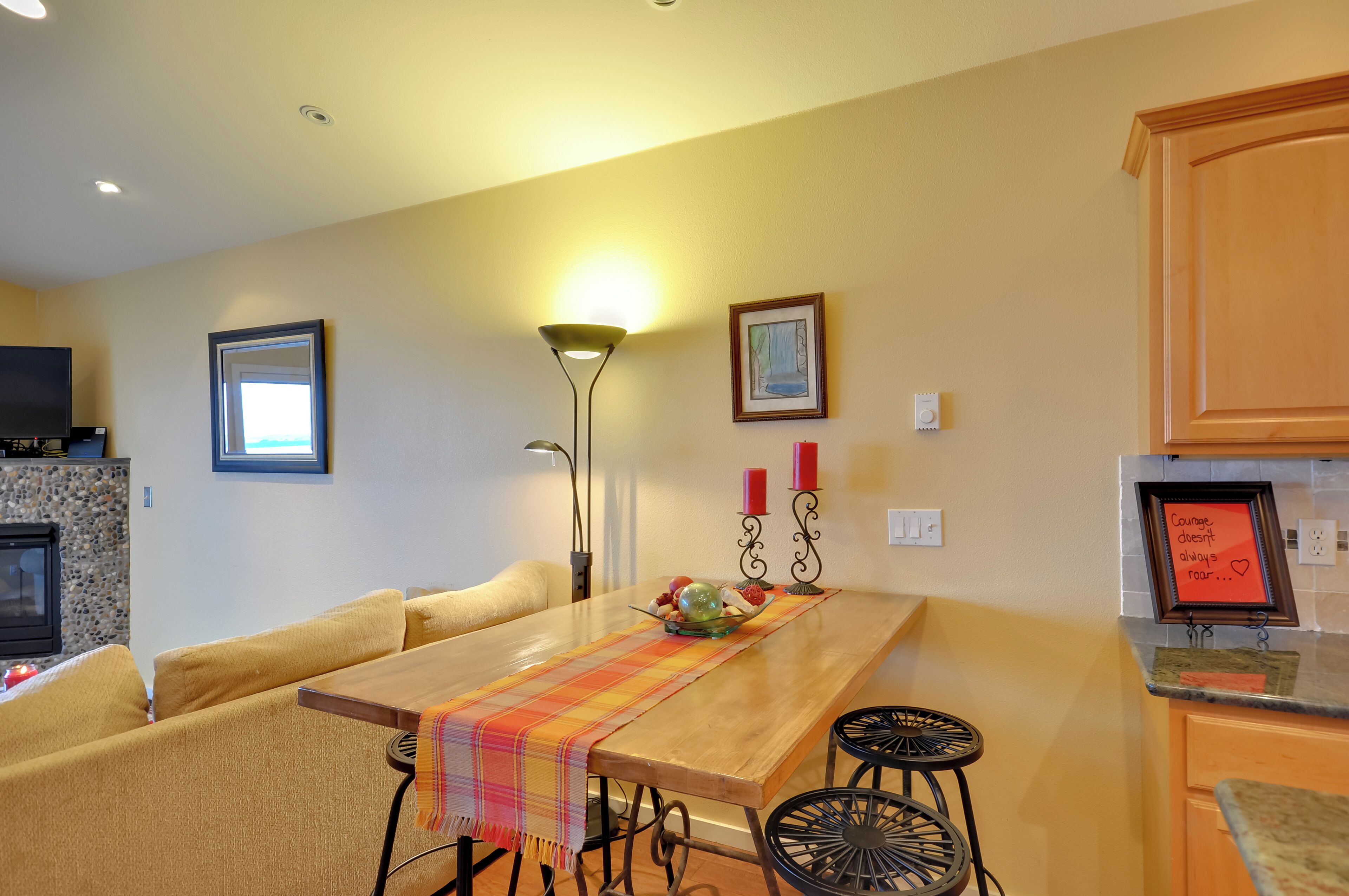 Un comedor con mesa de madera, sofá amarillo y sillas negras. Hay una chimenea, un cuadro enmarcado en la pared y una lámpara.