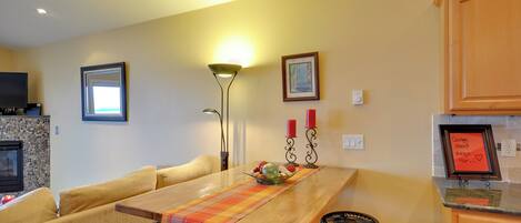 Un comedor con mesa de madera, sofá amarillo y sillas negras. Hay una chimenea, un cuadro enmarcado en la pared y una lámpara.