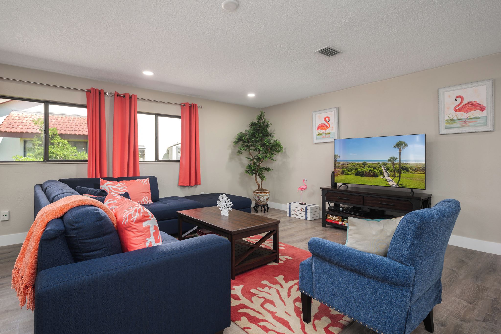 A living room with a sofa, armchair, coffee table, and TV. There is a plant, curtains, and artwork on the wall.