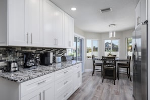 Una cocina moderna con armarios blancos, encimeras de granito y una zona de comedor con sillas y mesa de madera.