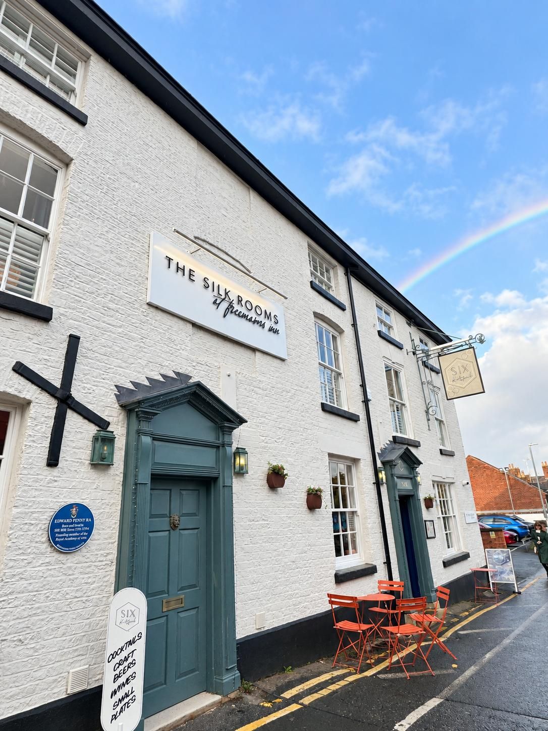 A building with a sign for 'The Six Rooms' pub, a traditional design with a green door, and outdoor seating with red chairs.