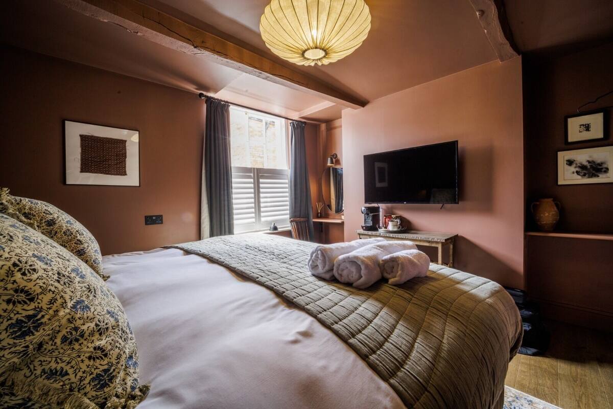 Ein Schlafzimmer mit einem großen Bett, einem Flachbildfernseher, einem Nachttisch mit Lampe und einem Fenster mit Vorhängen.