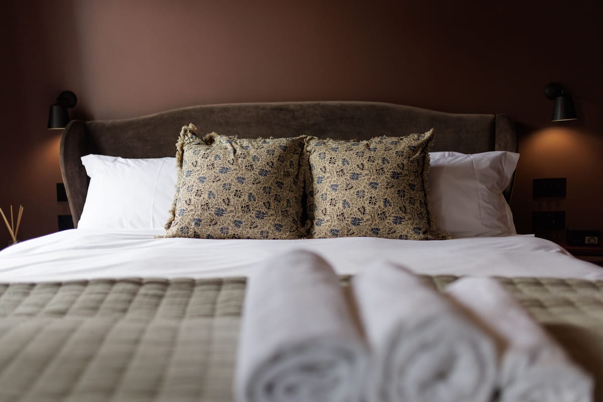 Ein ordentlich gemachtes Bett mit einer gesteppten Tagesdecke, zwei gemusterten Kissen und aufgerollten weißen Wäschestücken.