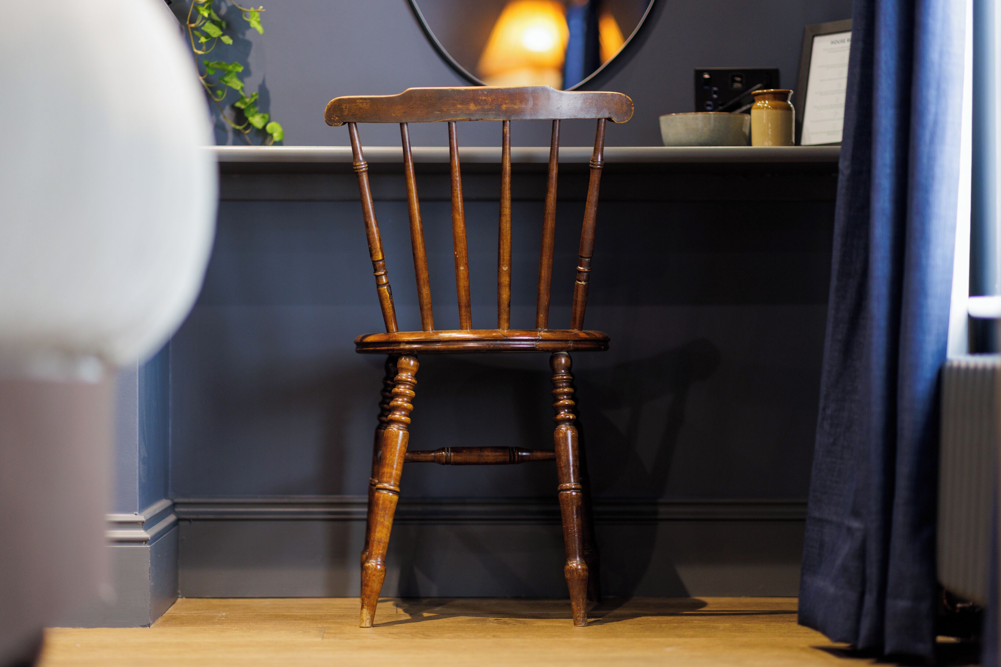 Una silla de madera con respaldo alto y patas torneadas, ubicada frente a una pared oscura con un estante encima.