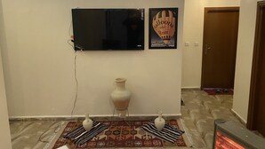 A room with a flat-screen TV mounted on the wall, a decorative poster, a white vase, a patterned rug, and a portable heater. - Nabati Hostel  (At-Taybeh)