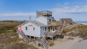 A beachfront house with a red door, a wooden deck, and a staircase leading to a second floor. - Amazing Beach house Steps from the Surf
"Nautical Endeavors" (Rodanthe)