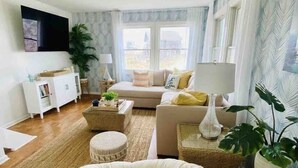A living room with a sofa, a round bean bag, a white cabinet, a TV, and a large window. - Amazing Beach house Steps from the Surf
"Nautical Endeavors" (Rodanthe)