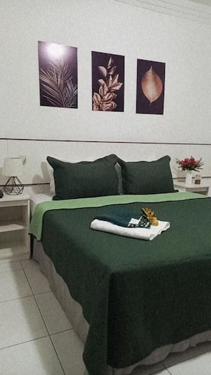 A neatly made bed with a green bedspread, two pillows, and a folded towel on the bed. There are three framed botanical prints on the wall above the bed. - Hotel Cuca Fresca (Cotia)