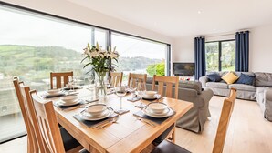 A dining area with a wooden table set for six, large windows offering a view of the countryside, and a grey sofa in the background. - Beach View (Ilfracombe)