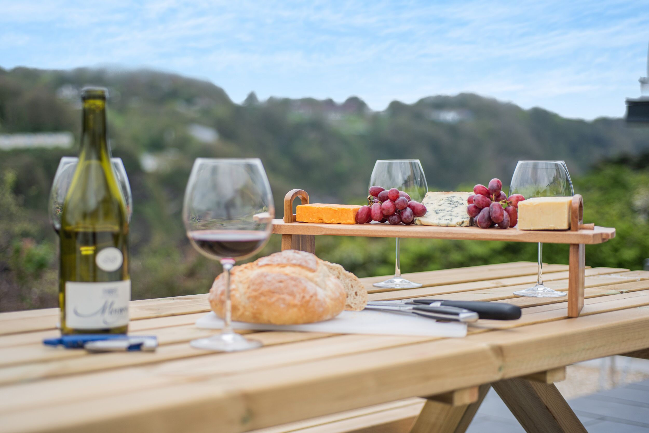 Un montaje para picnic con una botella de vino, copas, queso, uvas y hogaza de pan sobre una mesa de madera.