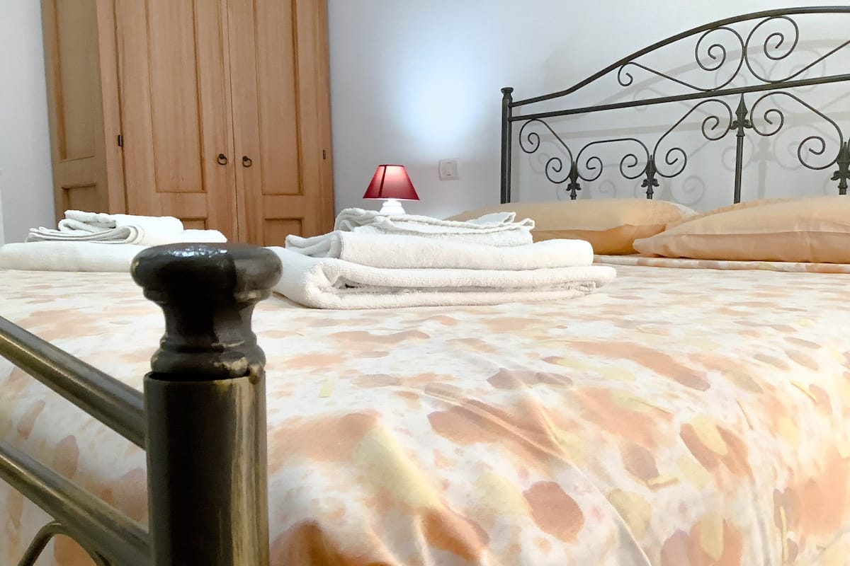 Uma cama com colcha estampada, uma luminária, toalhas dobradas e um guarda-roupa de madeira ao fundo.