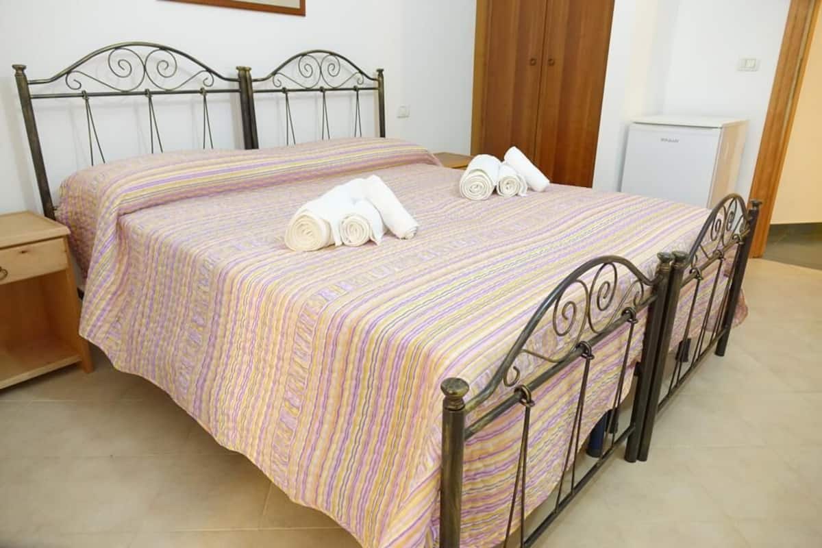 Uma cama bem arrumada com colcha listrada e toalhas dobradas, uma mesa de cabeceira de madeira, uma geladeira branca pequena e uma porta de madeira em um quarto de hotel.