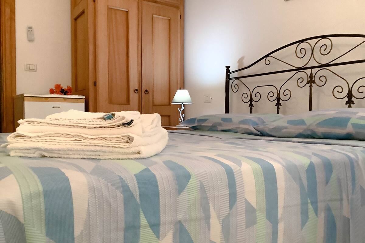 Uma cama bem arrumada com colcha listrada, uma pilha de toalhas dobradas, um abajur e um guarda-roupa de madeira em um quarto.